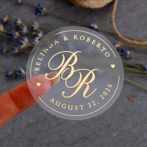 May include: A clear, round sticker with gold lettering. The text reads "Belinda & Roberto" around the top, with "August 22, 2026" at the bottom. The initials "BR" are in the center, with a heart symbol on each side.