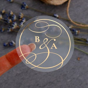 Op de afbeelding: Een transparante, ronde sticker met gouden letters en design. De sticker toont de letters "B & A" in een elegant lettertype, omgeven door decoratieve krullen. De sticker wordt door een vinger vastgehouden, met een vage achtergrond van lavendel en touw.