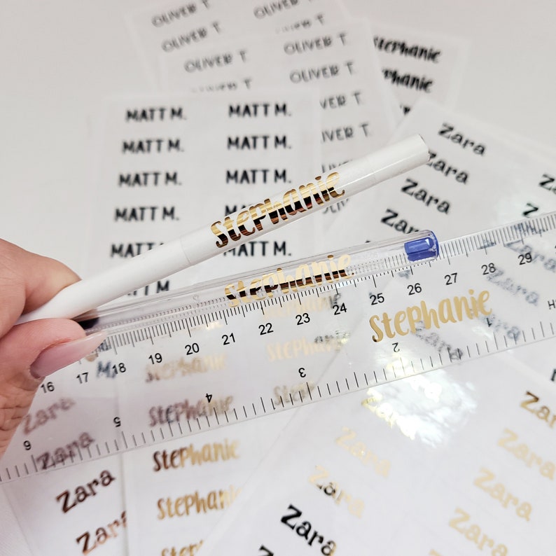 May include: A close-up of a white pen with gold lettering that says "Stephanie". The pen is lying on top of a clear ruler with black markings. The ruler is on top of a sheet of clear stickers with gold lettering. The stickers have the names "Zara", "Matt M.", "Stephanie", and "Oliver T" printed on them.