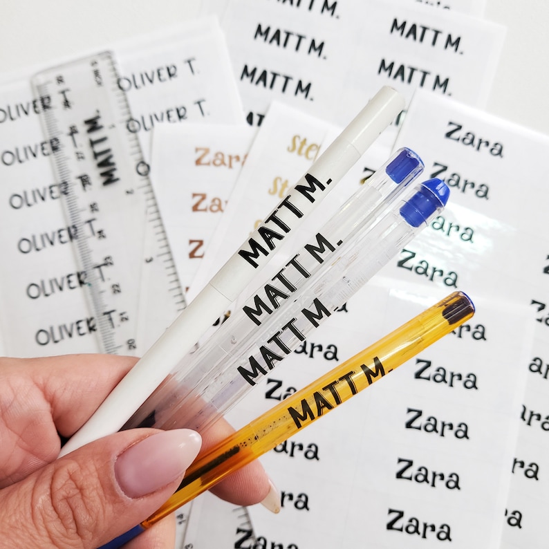 May include: Three pens with personalized labels that read "Matt M." and "Zara". The pens are white, clear, and orange. The labels are black and white.