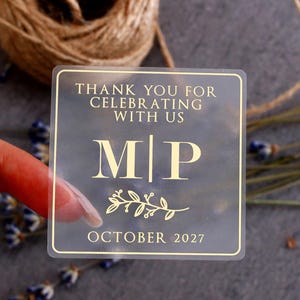 May include: A square, semi-transparent sticker featuring gold-colored text and a border. The text reads "THANK YOU FOR CELEBRATING WITH US", "M | P", and "OCTOBER 2027". A small gold-colored botanical illustration of a leafy branch is centered below the letters.