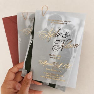 Può includere: Un invito di nozze in carta velina traslucida con una fotografia in bianco e nero di una coppia, impreziosito da scritte in lamina d'oro che riportano Adela & Nilson, 20 maggio 2023, ore 18:00, Marvel Golf Club, Santa Monica, CA, e un link.