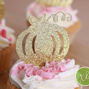 Peut inclure: D&eacute;cor de cupcake en forme de citrouille dor&eacute;e paillet&eacute;e avec un motif en spirale. Le d&eacute;cor est sur un cupcake avec du gla&ccedil;age blanc et des p&eacute;pites roses.