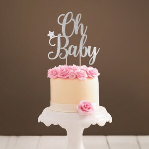 A cake topper made of silver glittered cardstock, spelling Oh Baby, decorated with butterflies. The font is hand-lettered typography.