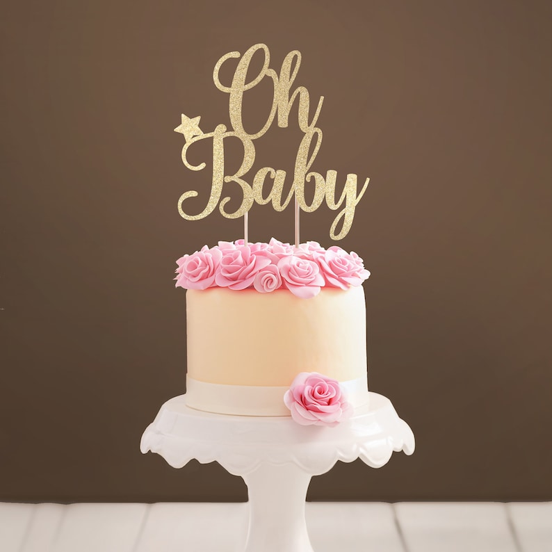 A cake topper made of gold glittered cardstock, spelling Oh Baby, decorated with butterflies. The font is hand-lettered typography.
