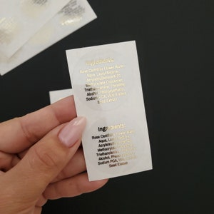 May include: Two round, white labels with gold lettering. The labels list the ingredients for a product. The ingredients include Rose Centifolia Flower Water, Aqua, Lauryl Betanie, Acrylates/Beheneth-25 Methacrylate Copolymer, Triethanolamine, Phenethyl Alcohol, Phenoxyethanol, Sodium PCA, Vitis Vinifera Seed Extract.