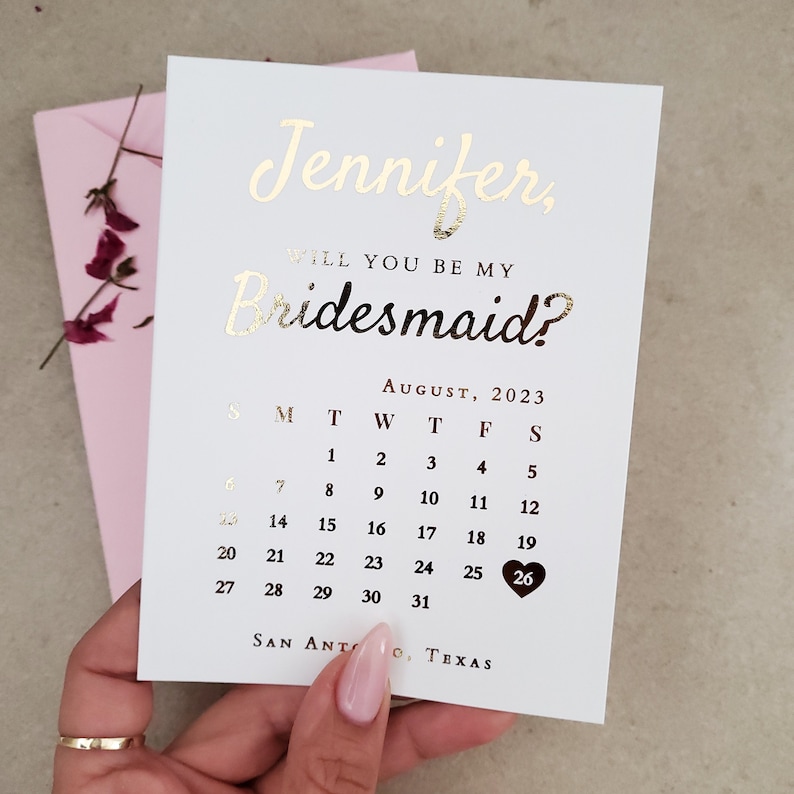 May include: A white card with gold foil lettering reading Jennifer, Will You Be My Bridesmaid? above an August 2023 calendar. The date 26 is highlighted with a dark heart. The card is positioned in front of a light pink envelope with dried flowers.