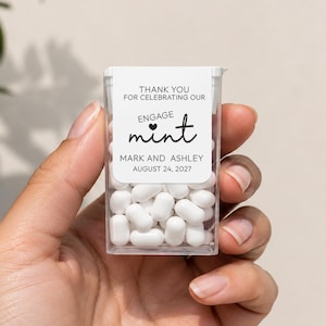 May include: A clear rectangular plastic container filled with white mints. A white label on the front reads: THANK YOU FOR CELEBRATING OUR ENGAGE mint MARK AND ASHLEY AUGUST 24, 2027. The text features a small heart icon above the word mint.