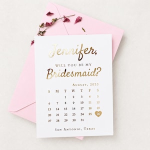 May include: A white card with gold foil lettering that reads Jennifer, Will You Be My Bridesmaid? above an August 2023 calendar with a gold heart over the 26th and San Antonio, Texas at the bottom, resting on a light pink envelope with dried flowers.