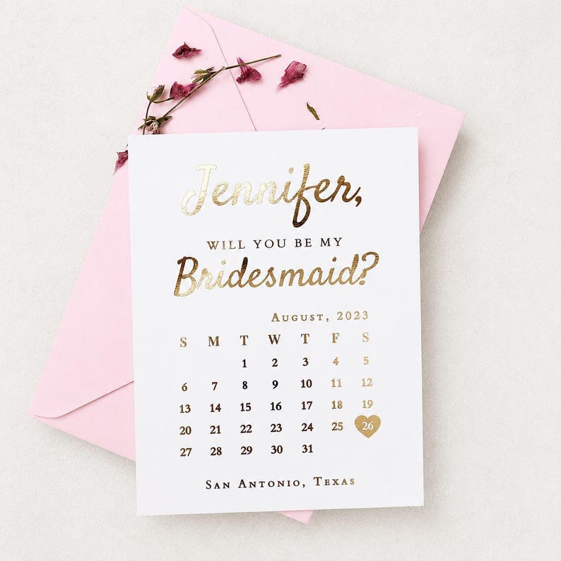 May include: A white card with gold foil lettering that reads Jennifer, Will You Be My Bridesmaid? above an August 2023 calendar with a gold heart over the 26th and San Antonio, Texas at the bottom, resting on a light pink envelope with dried flowers.