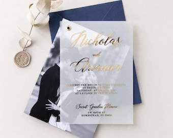Op de afbeelding: Een doorschijnende vellum trouwkaart met goudfolie belettering met de tekst Nicholas and Arianna, geplaatst over een zwart-witfoto van een koppel, vastgezet met een gouden oogje op een donkerblauwe envelop.