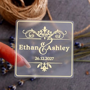 Può includere: Un adesivo quadrato semitrasparente con testo e decorazioni color oro. L'adesivo riporta i nomi Ethan e Ashley, una e commerciale e la data 26.12.2027, il tutto racchiuso all'interno di un sottile bordo color oro.