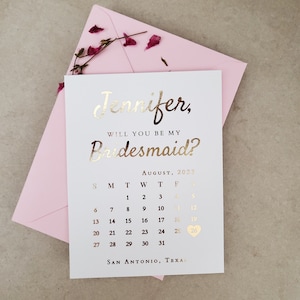 May include: A white card with metallic gold foil text reading Jennifer, Will You Be My Bridesmaid? above an August 2023 calendar with a heart icon on the 26th and San Antonio, Texas at the bottom, placed on a light pink envelope with dried petals.