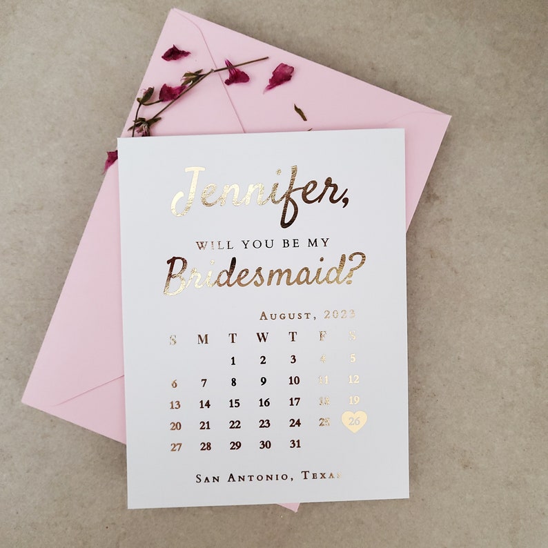 May include: A white card with metallic gold foil text reading Jennifer, Will You Be My Bridesmaid? above an August 2023 calendar with a heart icon on the 26th and San Antonio, Texas at the bottom, placed on a light pink envelope with dried petals.