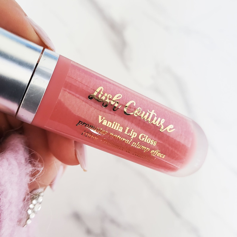 Puede incluir: Un tubo transparente de brillo de labios rosa con una etiqueta dorada que dice "Lush Couture Brillo de labios de vainilla promueve un efecto de volumen natural".