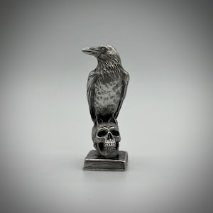 May include: A silver raven figurine perched atop a silver skull, both standing on a small silver base. The raven has a detailed, textured design and is positioned with its wings slightly spread.