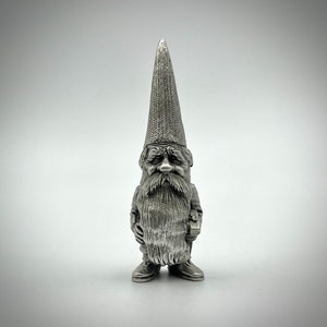 May include: A silver-colored gnome figurine with a long, pointed hat and a full beard. The gnome is wearing a jacket and boots, and has a stern expression. The figurine is made of metal and has intricate detailing.