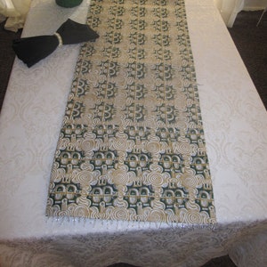 May include: A decorative table runner with a repeating pattern of green and gold shapes. The runner is placed on a white tablecloth and has a beaded fringe along the bottom edge. A black napkin with a silver ring sits on the table.