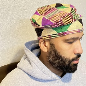 May include: A colorful, patterned hat with a brim. The hat features a geometric design in shades of purple, green, black, and yellow. The hat is made of woven fabric and has a rounded crown. The brim is a darker shade of the same colors.