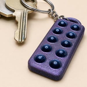 May include: A rectangular purple glitter keychain featuring ten dark blue circular indentations arranged in two parallel rows of five. The keychain is attached to a silver metal split ring that holds a set of brass-colored metal keys.