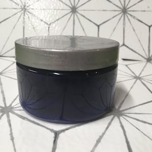 May include: A round, silver-lidded jar with a dark blue, patterned body. The jar is sitting on a white surface with a geometric pattern.