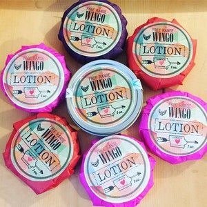 May include: A collection of Wingo Lotion containers, each wrapped in colorful tissue paper. The labels feature the text "Free Range Wingo Lotion" with an arrow and heart design, and the text "Made with in Iowa, 1 oz."