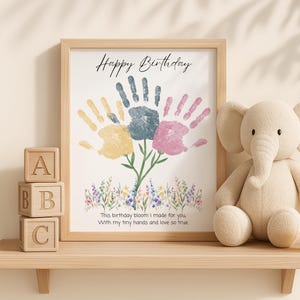 Puede incluir: Una impresión enmarcada con un diseño de flores de huellas de manos, las palabras "Happy Birthday" y un dulce mensaje. Junto a ella, un elefante de peluche y bloques de madera que deletrean "ABC" se encuentran en un estante. Un regalo encantador.