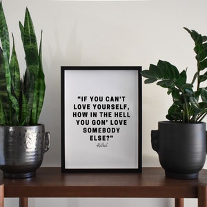 May include: A black framed print with a white background and black text that reads "IF YOU CAN'T LOVE YOURSELF, HOW IN THE HELL YOU GON' LOVE SOMEBODY ELSE?" -Rusbaul. The print is sitting on a wooden shelf between two black potted plants.
