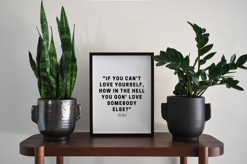 May include: A black framed print with a white background and black text that reads "IF YOU CAN'T LOVE YOURSELF, HOW IN THE HELL YOU GON' LOVE SOMEBODY ELSE?" -Rusbaul. The print is sitting on a wooden shelf between two black potted plants.