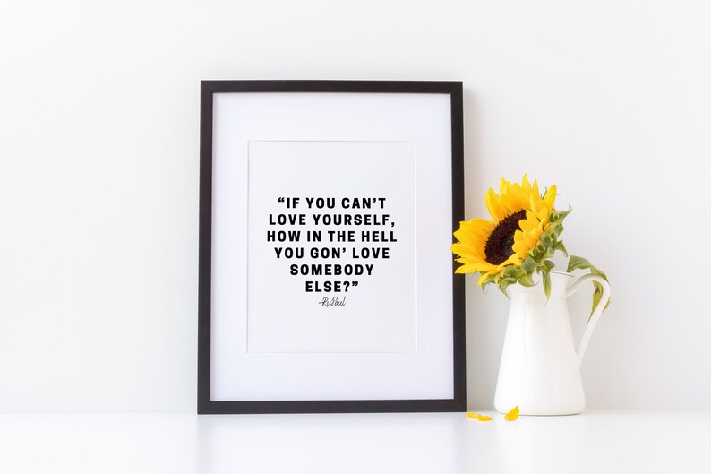 May include: A black framed print with a white background and a quote in black text: "IF YOU CAN'T LOVE YOURSELF, HOW IN THE HELL YOU GON' LOVE SOMEBODY ELSE?" -  Rural. A sunflower in a white pitcher sits on a white surface below the print.