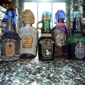 May include: Five decorative potion bottles with colorful wax drips and labels. The bottles are labeled "Spider Web", "Bone Powder", "Witch's Brew", "White Wolf", and "Death Potion". The bottles have a vintage, gothic aesthetic.