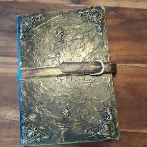 Peut inclure: Un journal artisanal avec une couverture texturée, dorée foncée et des coins décoratifs. Le journal est doté d'une sangle en cuir avec une boucle argentée pour le maintenir fermé. La couverture a un aspect vieilli, lui donnant une esthétique vintage.