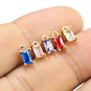 Könnte beinhalten: Sechs goldene Charms mit rechteckigen Edelsteinen in verschiedenen Farben. Die Charms haben eine Öse oben zum Anbringen an einer Halskette oder einem Armband.