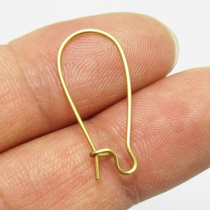 Op de afbeelding: Een goudkleurige oorhaak, vastgehouden tussen vingers, tegen een witte achtergrond. De haak heeft een langwerpige ovale vorm met een kleine, rechte pin aan de onderkant om de oorbel te bevestigen.