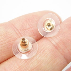 May include: Two clear plastic earring backs with gold-colored metal posts. The backs are round and have a slightly raised center.