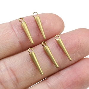 Puede incluir: Cinco colgantes de metal de color dorado en forma de cono con un bucle en la parte superior. Los colgantes se muestran en una mano.