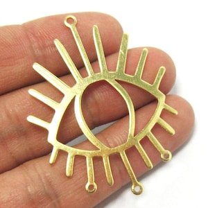 Könnte beinhalten: Ein goldfarbener Metallanhänger in Augenform mit Sonnenstrahlendesign. Der Anhänger hat eine ovale Augenform in der Mitte, umgeben von radialen Linien und kleinen Schlaufen zur Befestigung an anderen Schmuckkomponenten.