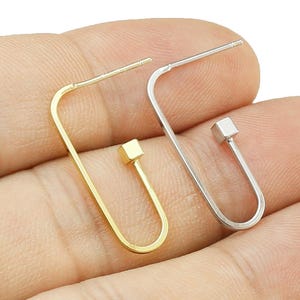 Op de afbeelding: Twee minimalistische oorbellen, één goudkleurig en één zilverkleurig, elk met een geometrisch ontwerp. De oorbellen hebben een rechthoekige vorm met een klein vierkant accent aan de onderkant. De pin is recht en loopt naar boven.