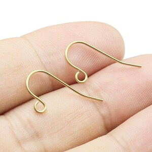 May include: Two gold-toned earring hooks, also known as ear wires, are shown on a hand. The hooks are made of metal and have a simple, classic design.