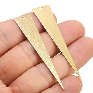 Puede incluir: Dos formas triangulares de metal de color dorado con un agujero en la parte superior. Los triángulos están cepillados y tienen un acabado mate.