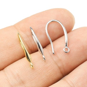 Puede incluir: Tres ganchos para pendientes, uno dorado y dos plateados, se muestran en una mano. Los ganchos están diseñados para sujetar pendientes.