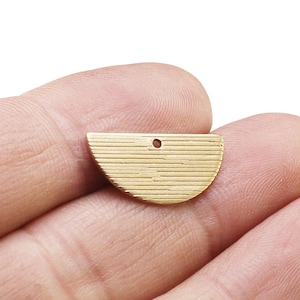 May include: A gold-coloured, half-circle pendant with a textured, horizontal-line pattern. The pendant has a small hole at the top for stringing. The pendant is held in a person's hand.