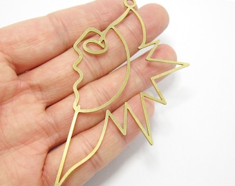 May include: A gold-colored metal pendant with a geometric design. The pendant features a stylized face with a pointed chin and a jagged, sunburst-like shape on the right side.