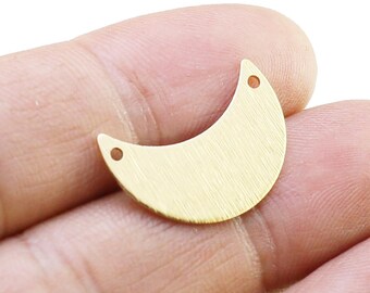 Puede incluir: Un dije de media luna de color dorado con dos pequeños agujeros en la parte superior. El dije tiene un acabado de metal cepillado y está sostenido en la mano de una persona.