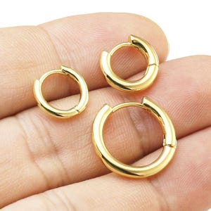 Könnte beinhalten: Drei goldfarbene Creolen in verschiedenen Größen. Die Ohrringe sind rund und haben eine glänzende Oberfläche. Die Reifen werden in einer Hand vor weißem Hintergrund gehalten.