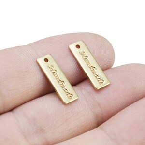 以下が含まれることがあります： 「Handmade」と刻印された金色の金属タグが2つ。タグの上部に穴が開いており、製品に取り付けることができます。