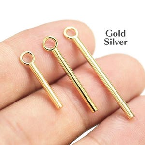 Puede incluir: Tres hallazgos de joyería dorados con un lazo en la parte superior. Los hallazgos son de diferentes longitudes y se sostienen en una mano. El texto "Gold Silver" está en el fondo.