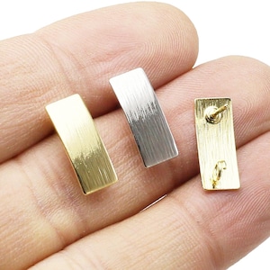 May include: Two pairs of earrings, one gold and one silver, with a rectangular shape and a brushed finish. The earrings are shown on a hand.