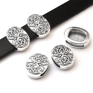 May include: Silver-coloured oval metal clasps with an ornate, swirling design. Some clasps are attached to a black leather strap. The clasps are displayed on a white background. The clasps are likely for jewellery or leathercraft projects.