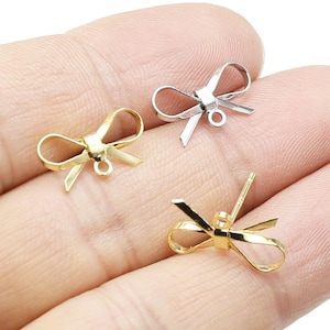 Könnte beinhalten: Drei goldfarbene Metall-Schleifen-Anhänger mit einer Öse zum Anbringen an Schmuck oder anderen Bastelarbeiten. Die Schleifen sind in verschiedenen Größen und werden auf einer Hand gezeigt.